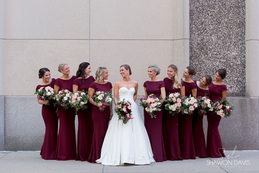Bride in white gown with bridesmaids in burgundy dresses holding bouquets against a stone wall