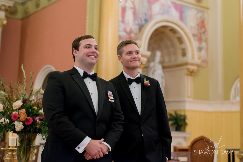 Two men in black tuxedos standing together in a church, smiling during a wedding ceremony.