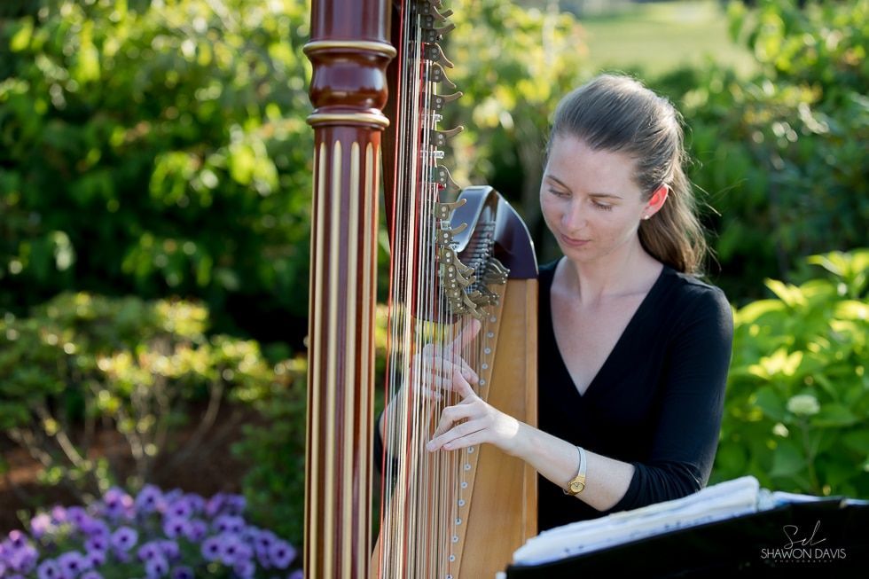 Woman playing a harp outdoors in a garden, with flowers and greenery in the background
