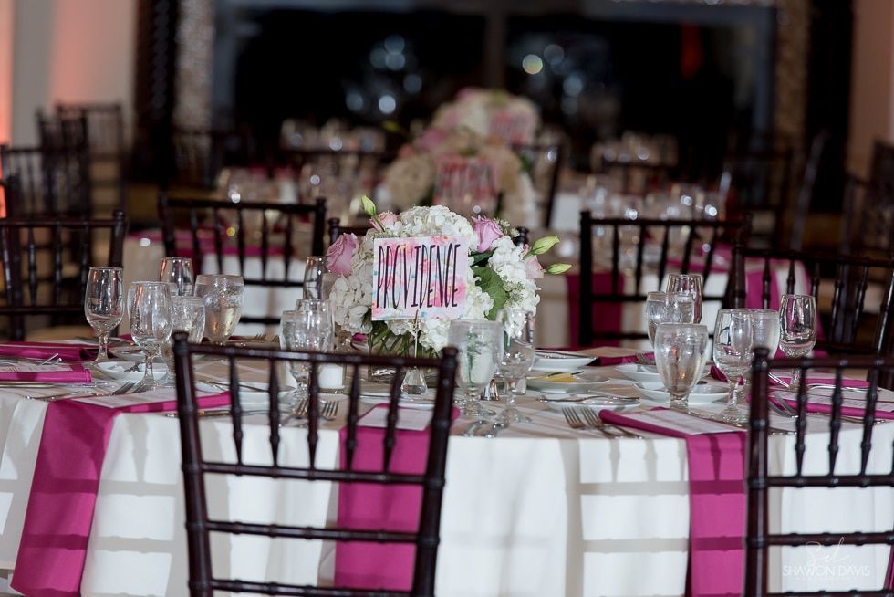 Decorated banquet table with white linens, pink accents, glassware, and floral centerpiece at an event