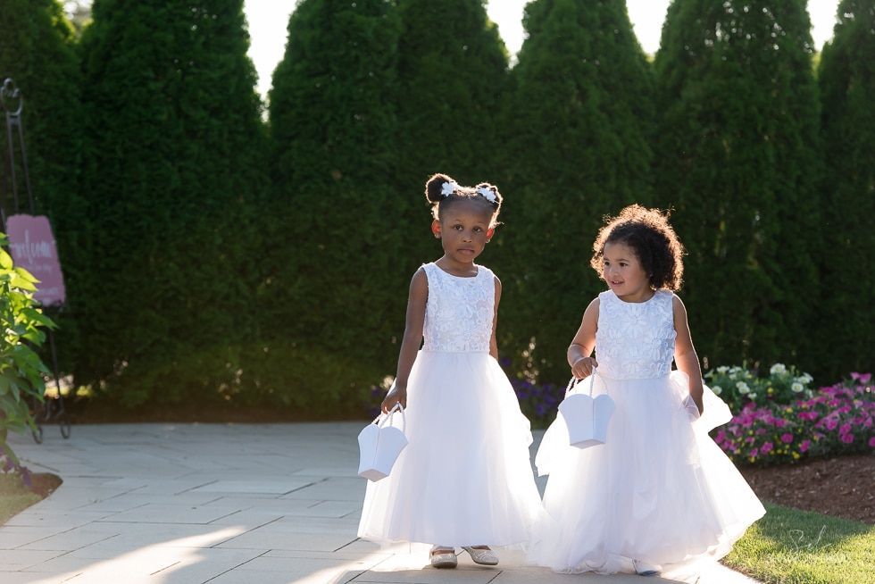 Two children in white dresses holding baskets, walking on a sunlit path in front of tall green hedges.