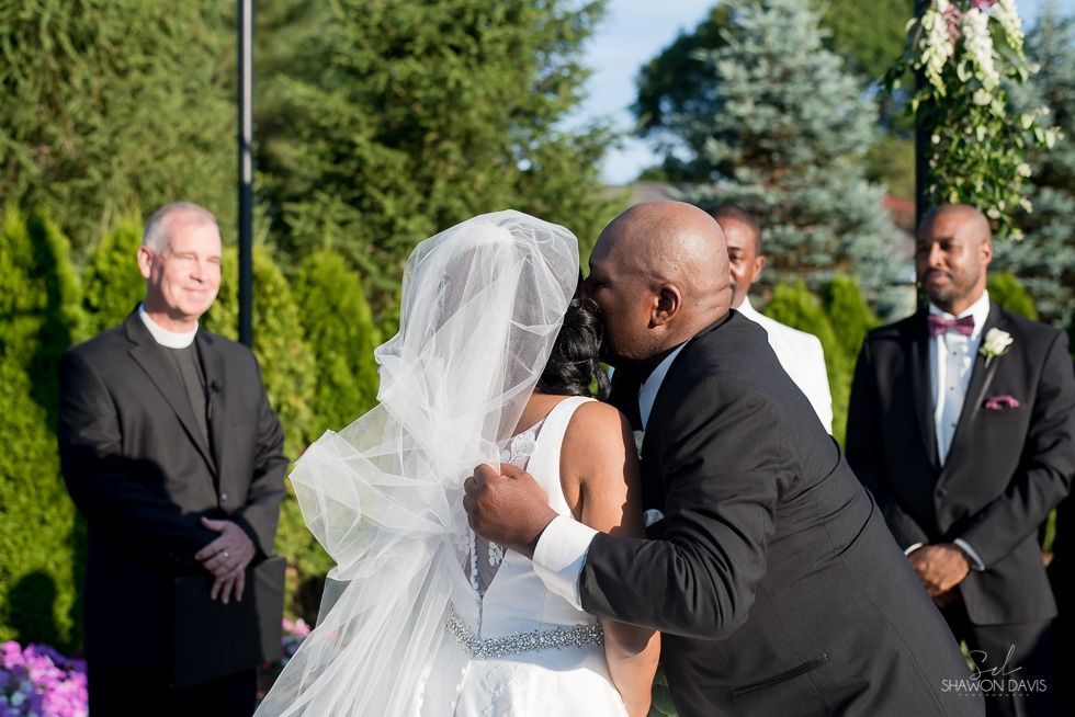 Bride and groom kiss during outdoor wedding ceremony, with officiant and groomsman nearby.