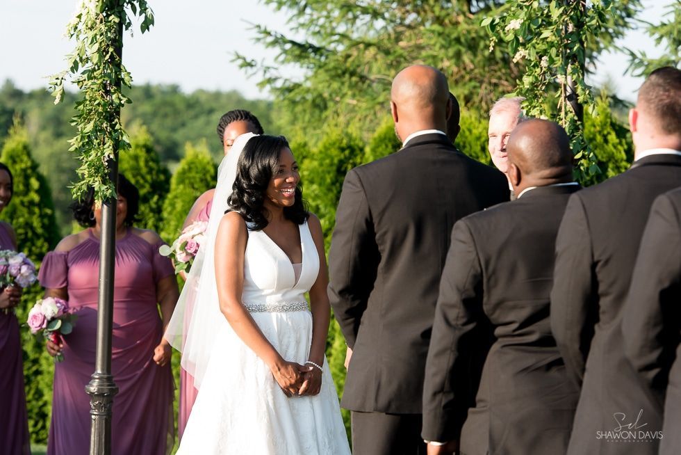 Bride in white dress smiles at outdoor wedding ceremony with guests in formal attire