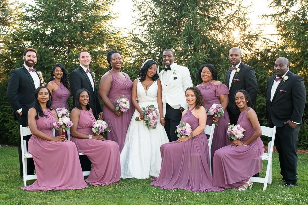 Wedding party posing outdoors, with bride and groom centered among attendants in lavender dresses and dark suits.