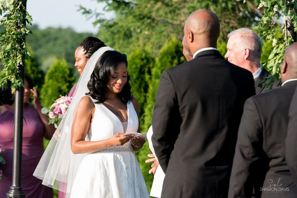 Bride reading vows outdoors at a wedding ceremony, with guests and greenery in the background
