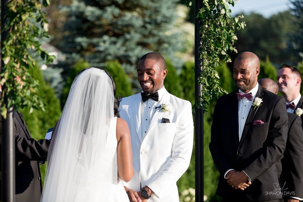 Bride and groom exchange vows outdoors, with guests watching in the background.