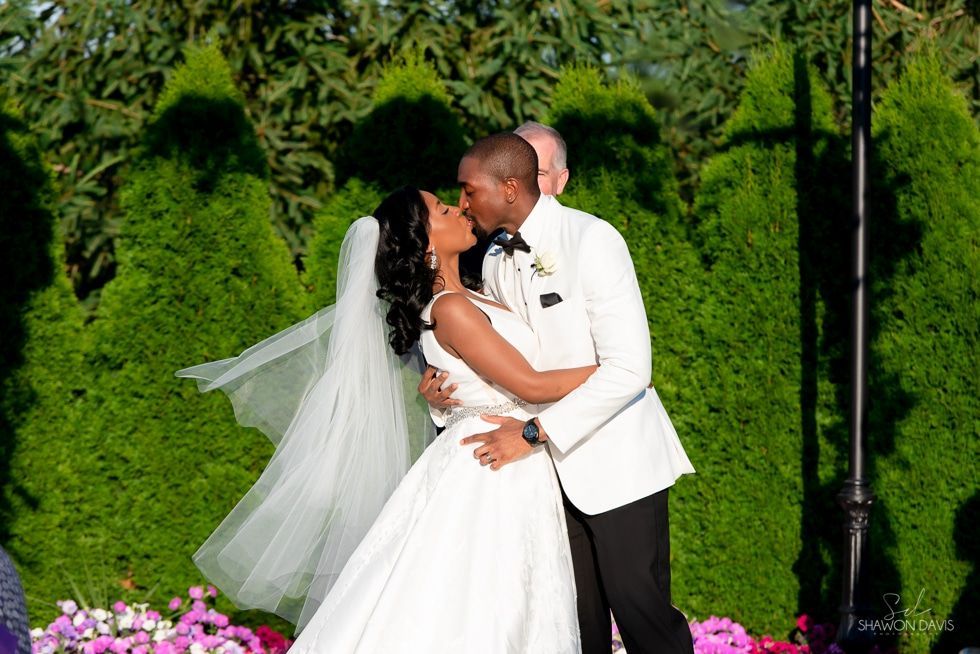Bride and groom kiss in white wedding attire before a green hedge backdrop.