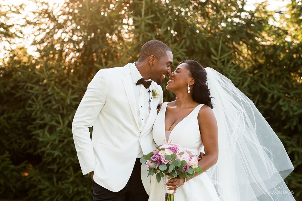 Bride and groom in white wedding attire sharing a smile outdoors, bride holding a bouquet