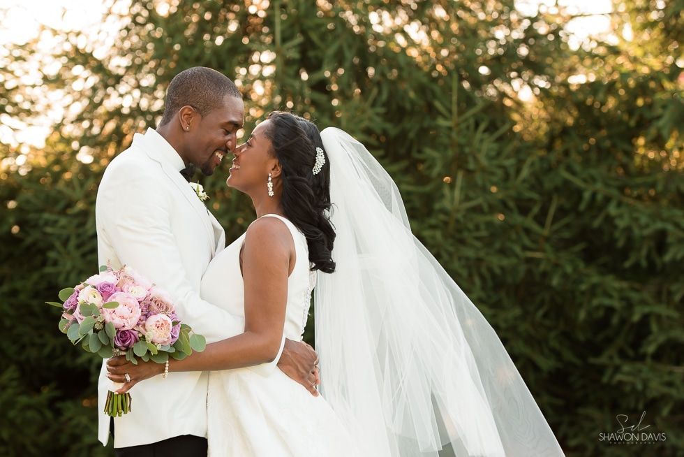 Bride and groom embracing outdoors, holding a bouquet, with a flowing white veil and greenery behind them