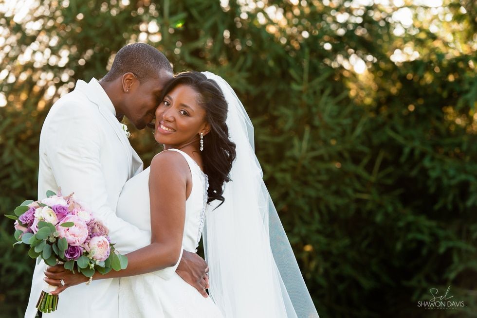 Couple embracing outdoors in wedding attire, with a bouquet and greenery in the background.