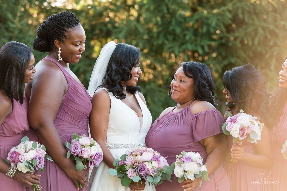 Five bridesmaids in purple dresses hold pink bouquets outdoors, smiling together.
