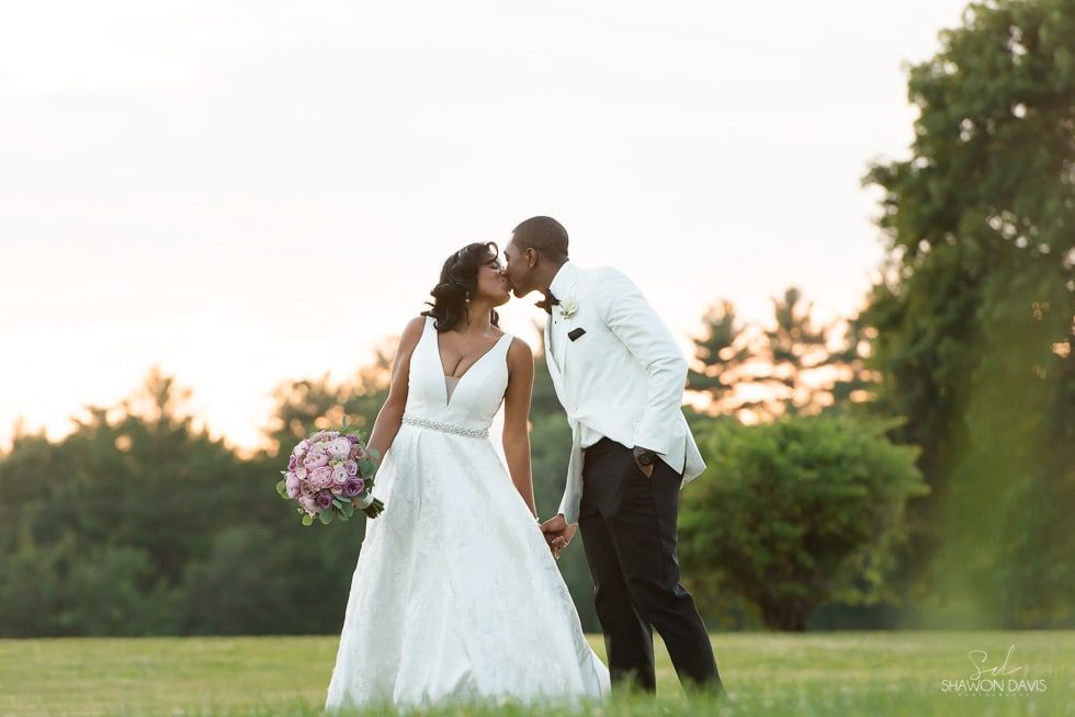 Bride and groom kiss in a grassy field at sunset, bride holding a bouquet of pink flowers.