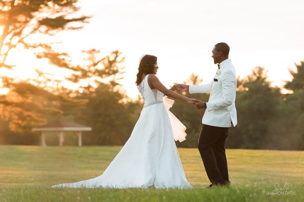 Bride in a white gown and groom in a white jacket holding hands at sunset in a grassy field