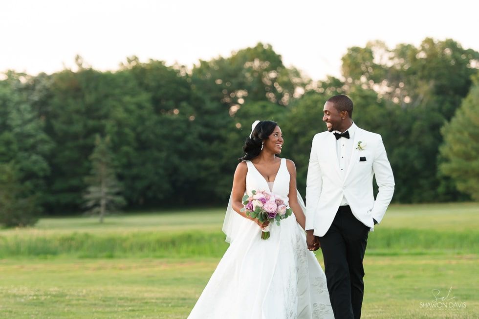 Bride and groom in white wedding attire walking hand in hand on a grassy field