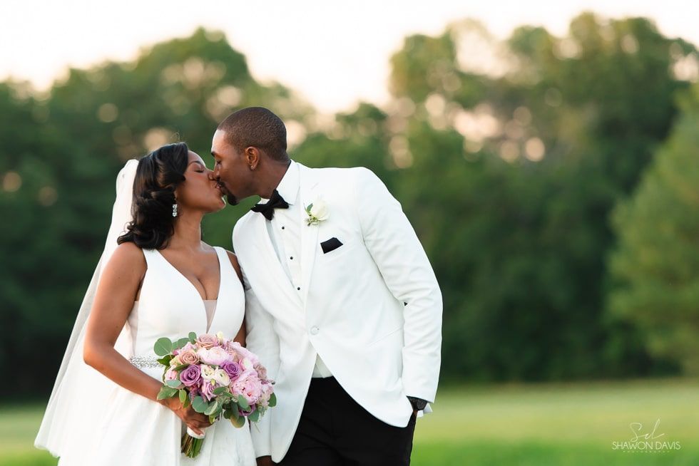 Newlyweds kiss outdoors in a green field, bride holding a bouquet and groom in a white tuxedo
