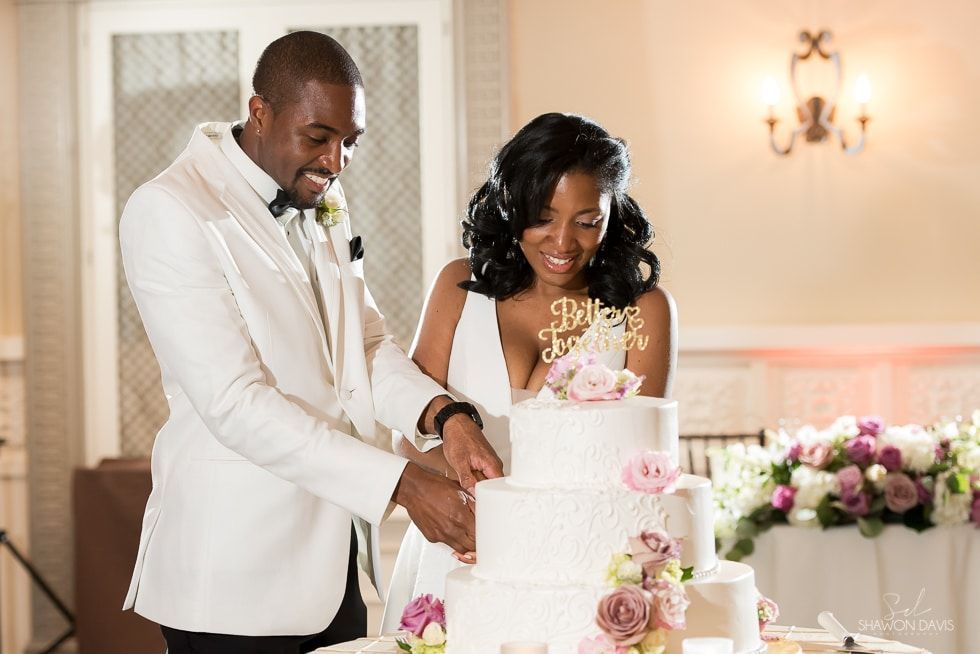 Couple in formal wear cutting a white wedding cake with pink flowers at a reception