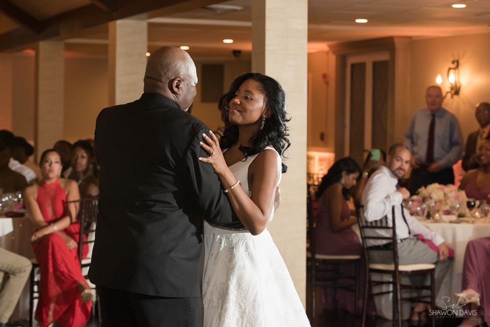 Couple slow dancing at a wedding reception, with guests seated at tables in the background.