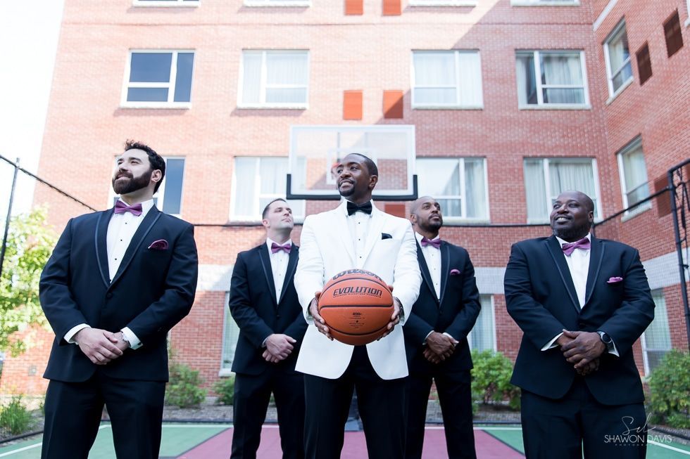 Five men in suits stand outdoors in front of a brick building, one holding a basketball.