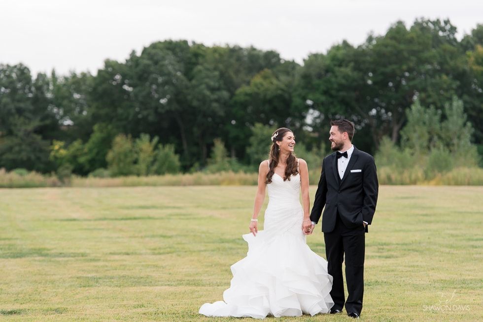 Bride and groom walking hand in hand across a grassy field, with trees in the background.