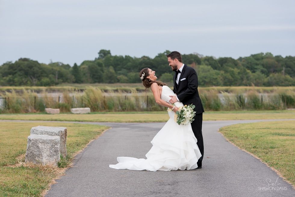 Bride and groom kissing outdoors on a paved path beside a grassy field