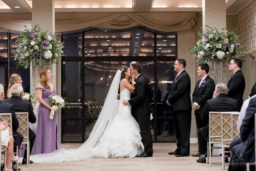 Bride and groom kiss at indoor wedding ceremony, flanked by attendants and floral arrangements.