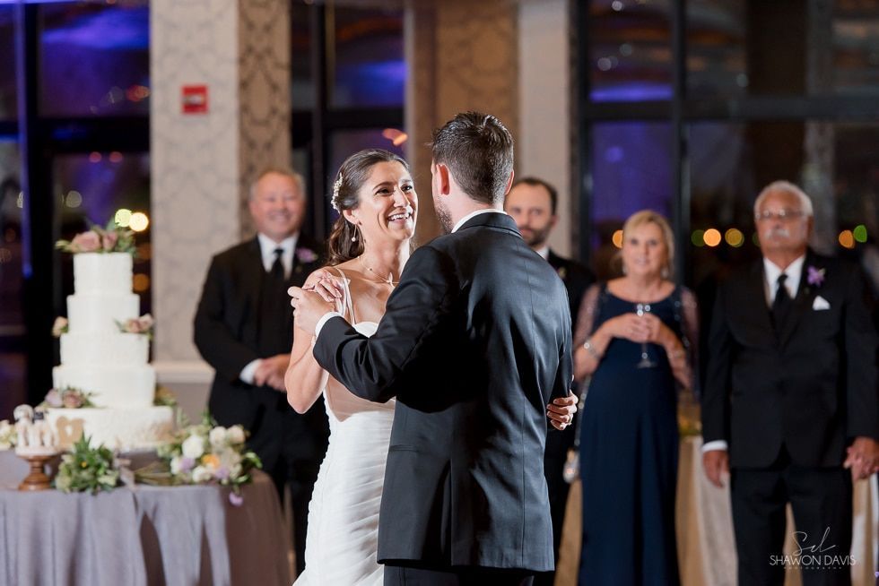 Bride and groom dancing at a wedding reception with guests and a tiered cake in the background