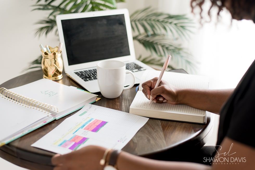 Person writing at a desk with an open laptop, notebook, coffee mug, and charts in a bright workspace