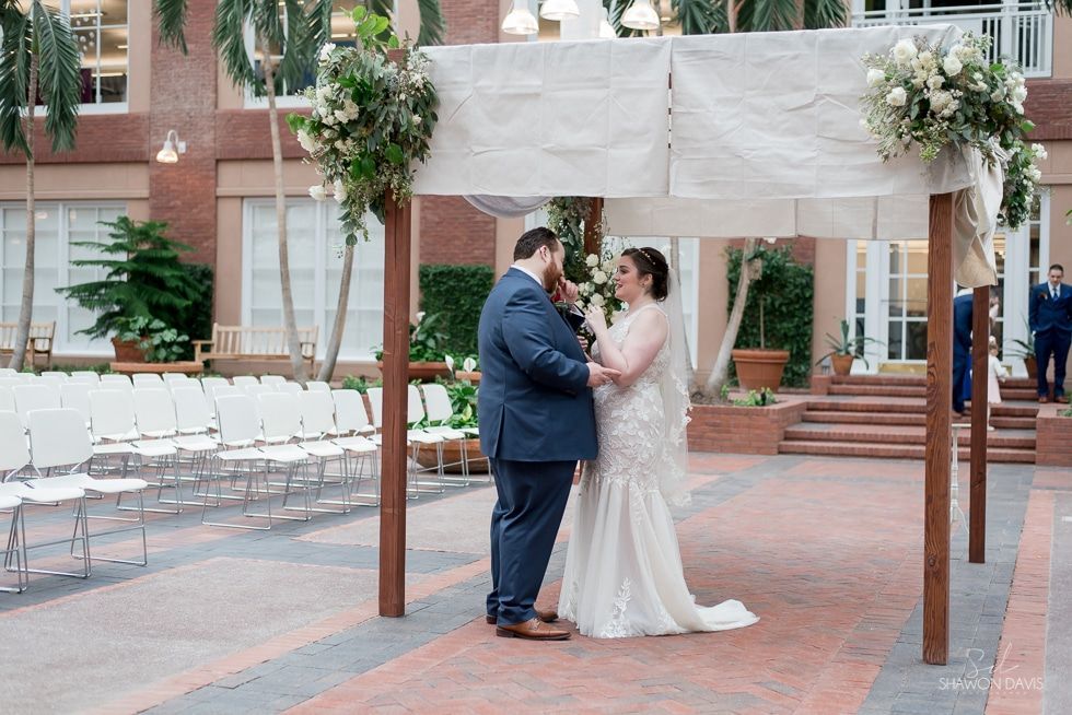 Couple in wedding attire exchanging vows under a floral canopy in an outdoor courtyard
