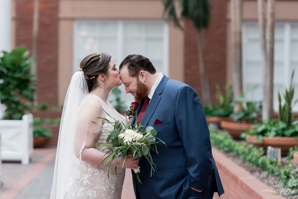 Bride and groom touch foreheads in a garden courtyard, holding a bouquet and wearing wedding attire