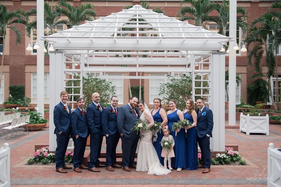 Wedding party posing under white gazebo in a garden courtyard, bride in white and bridesmaids in blue bouquets