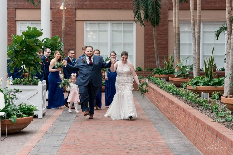 Wedding party walking down a brick courtyard, bride in white dress and groom in navy suit leading.