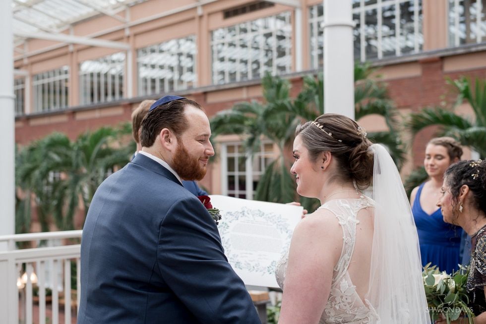 Bride and groom face each other during an outdoor wedding ceremony, with guests in the background.