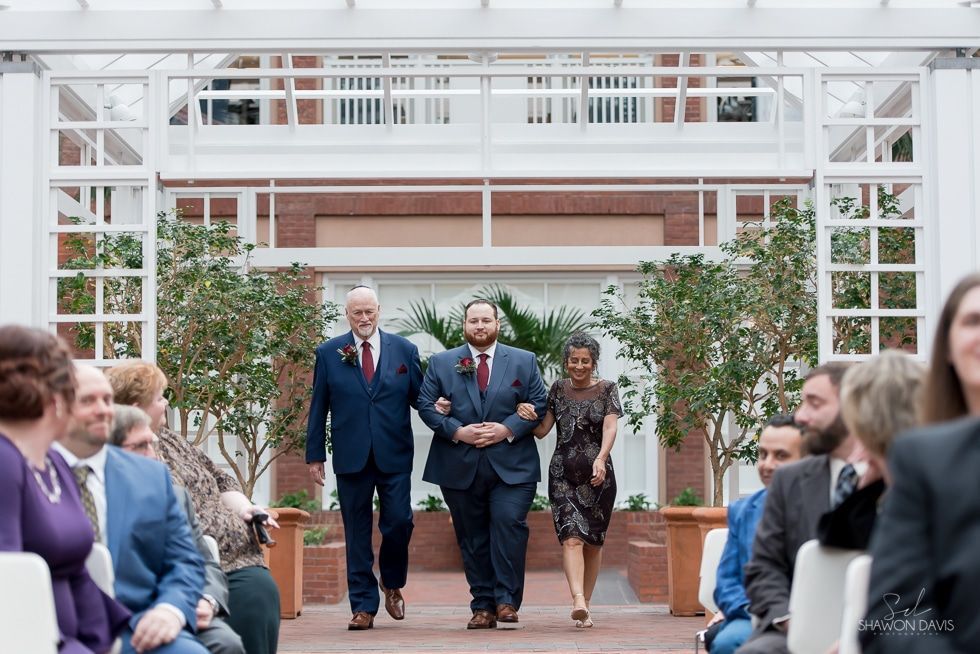 Three people walking down an aisle under a white pergola at an outdoor ceremony