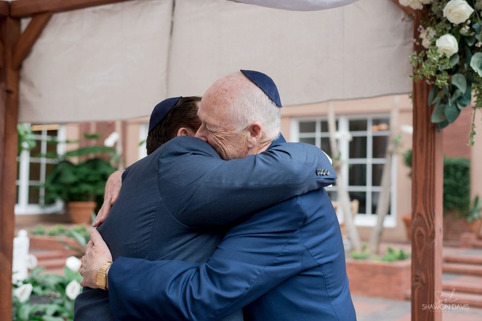 Two people in blue suits embracing under a decorated outdoor archway