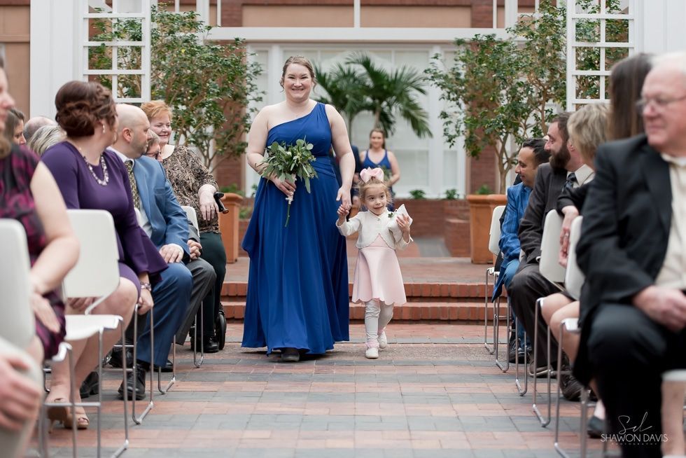 Woman in a royal blue dress walks down an aisle holding flowers, with two children beside her at an outdoor ceremony.