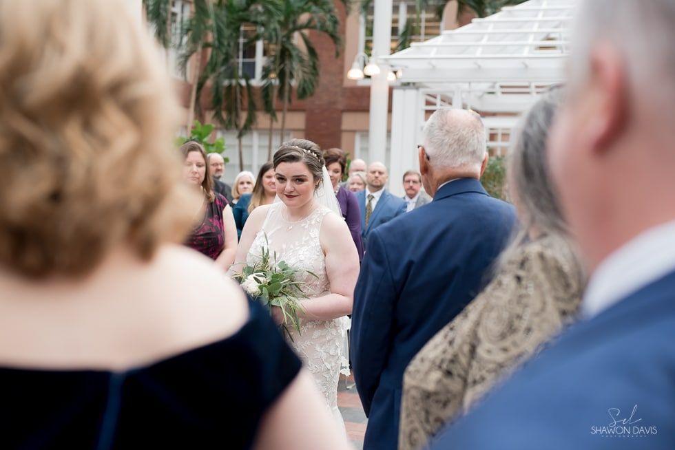 Bride walking through an outdoor wedding crowd, holding a bouquet and smiling beside a man in a navy suit.