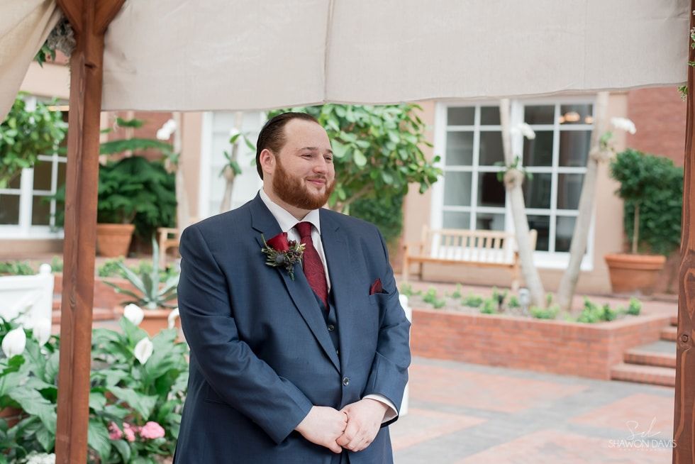 Man in navy suit with red tie stands smiling under a canopy in a brick courtyard