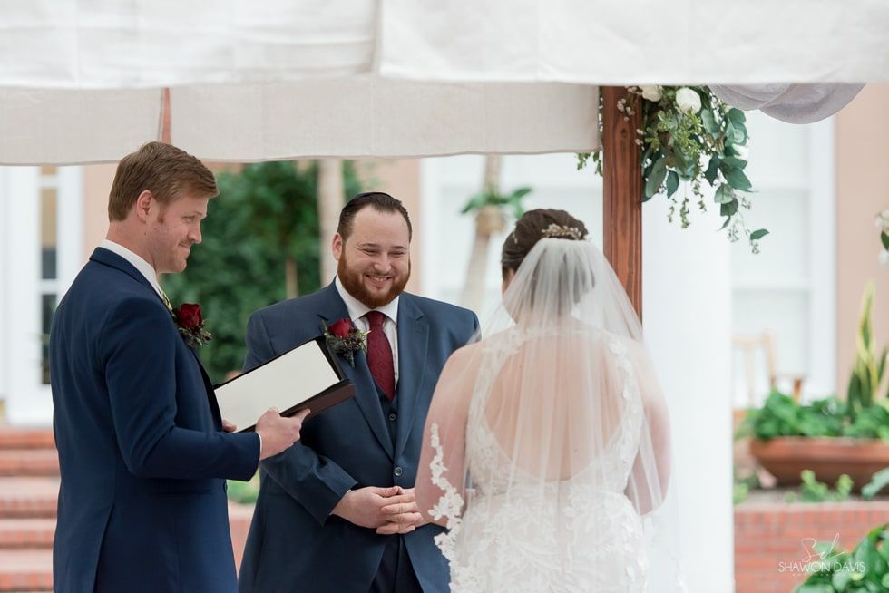 Wedding ceremony under a white canopy; officiant reads vows as couple holds hands.