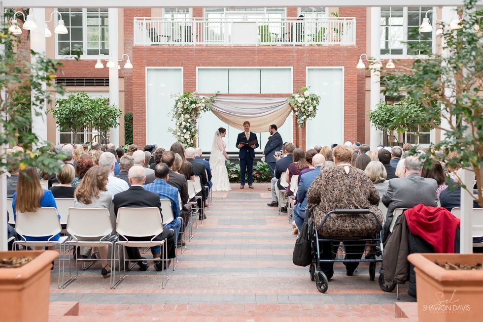Outdoor wedding ceremony in a courtyard, with guests seated facing an altar under white floral draping.