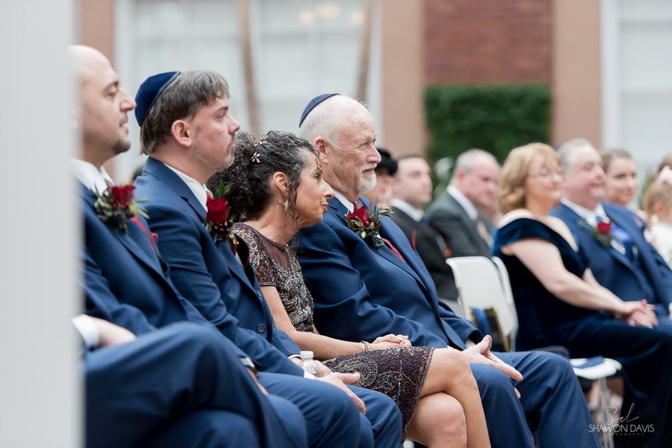People seated in formal attire at an outdoor ceremony, facing forward in a row of chairs.