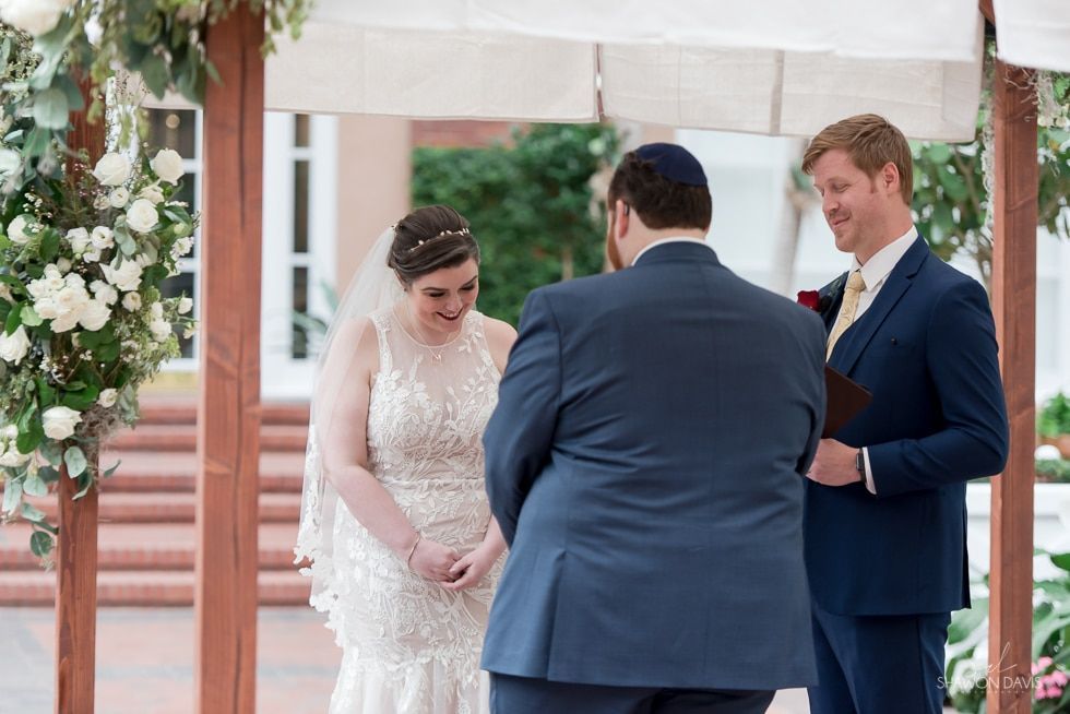 Wedding ceremony under a white canopy, bride and groom standing with an officiant beside floral arrangements