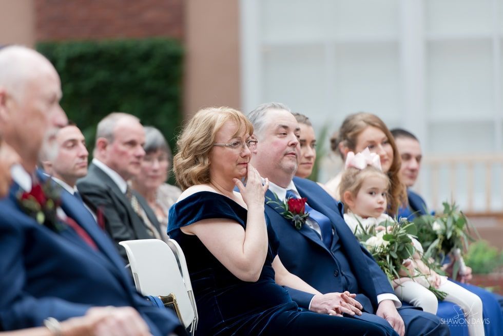 Seated guests at a formal ceremony, one woman holding a tissue with an emotional expression