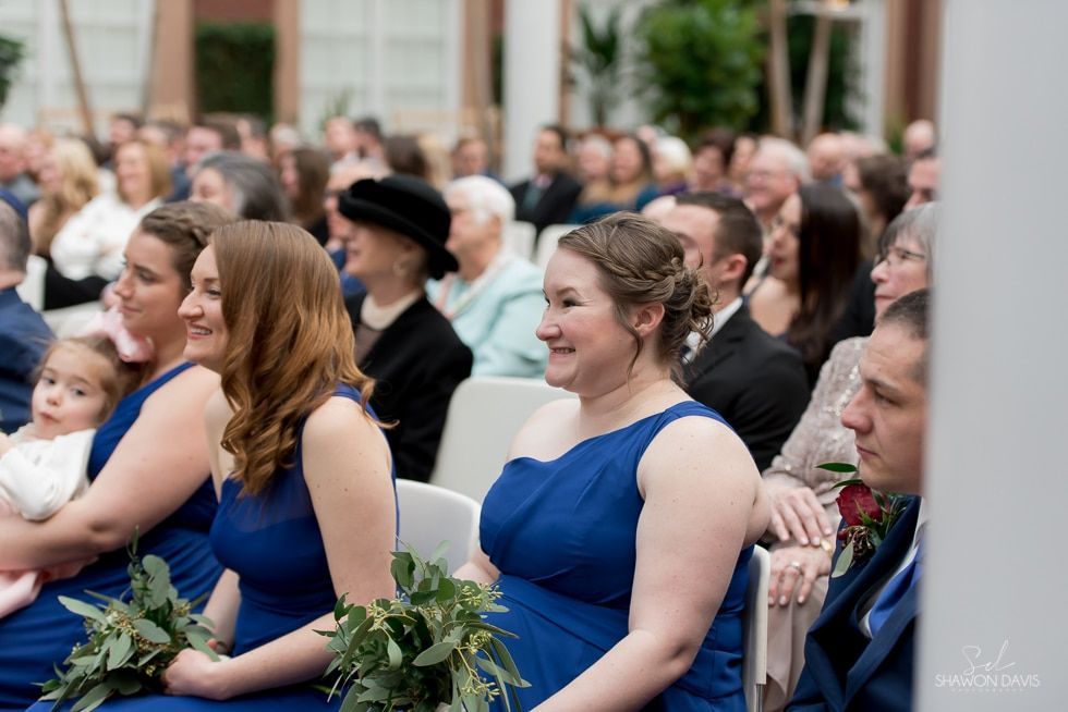 Guests in blue dresses seated at an outdoor ceremony, smiling and holding greenery.