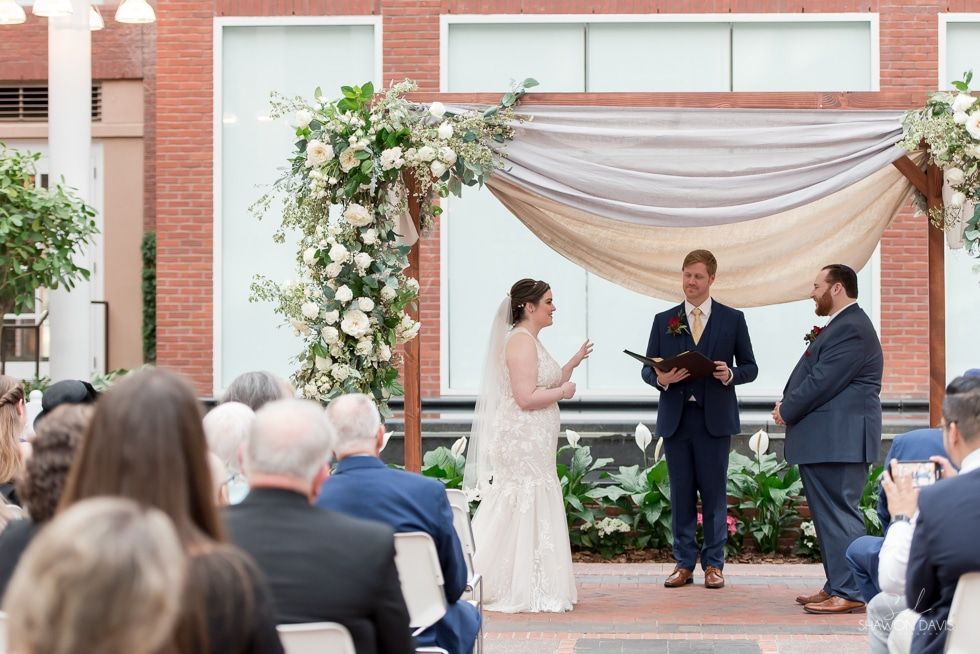 Wedding ceremony under a floral arch, with an officiant, bride, groom, and seated guests outdoors