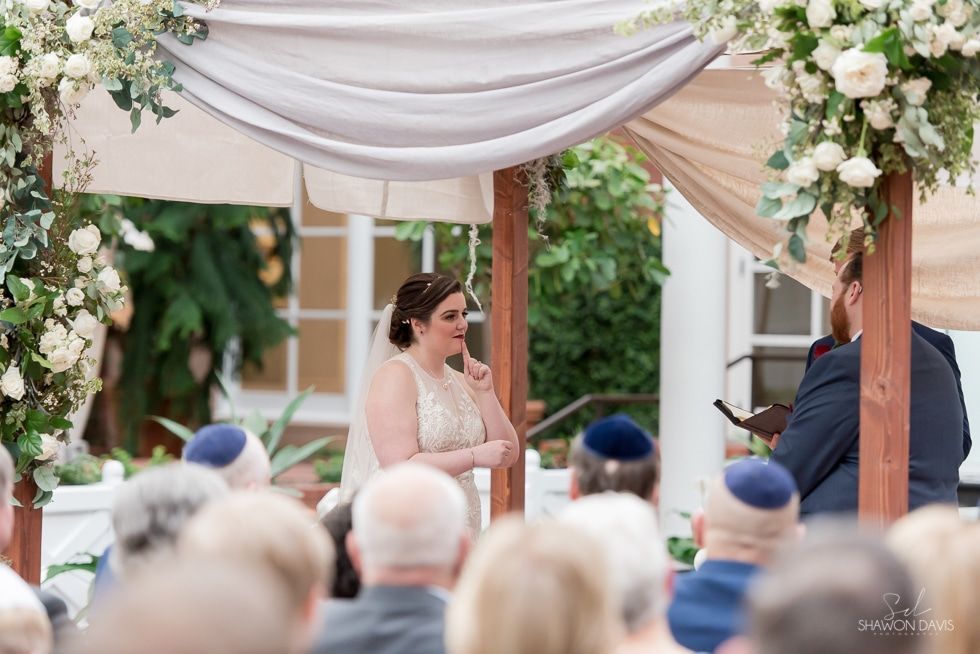 Bride standing under floral arch during outdoor wedding ceremony, facing seated guests
