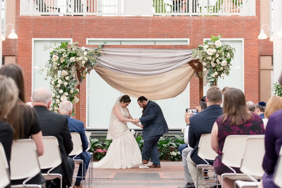 Wedding ceremony under floral arch, bride and groom holding hands outdoors, guests seated nearby