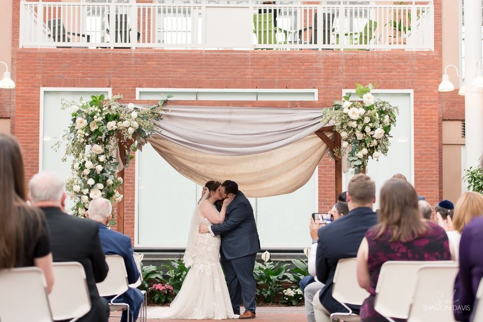 Bride and groom kissing under floral arch at outdoor wedding ceremony in front of brick building
