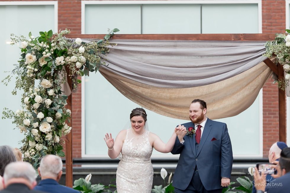 Wedding couple leaving ceremony under floral arch, smiling and holding hands outdoors
