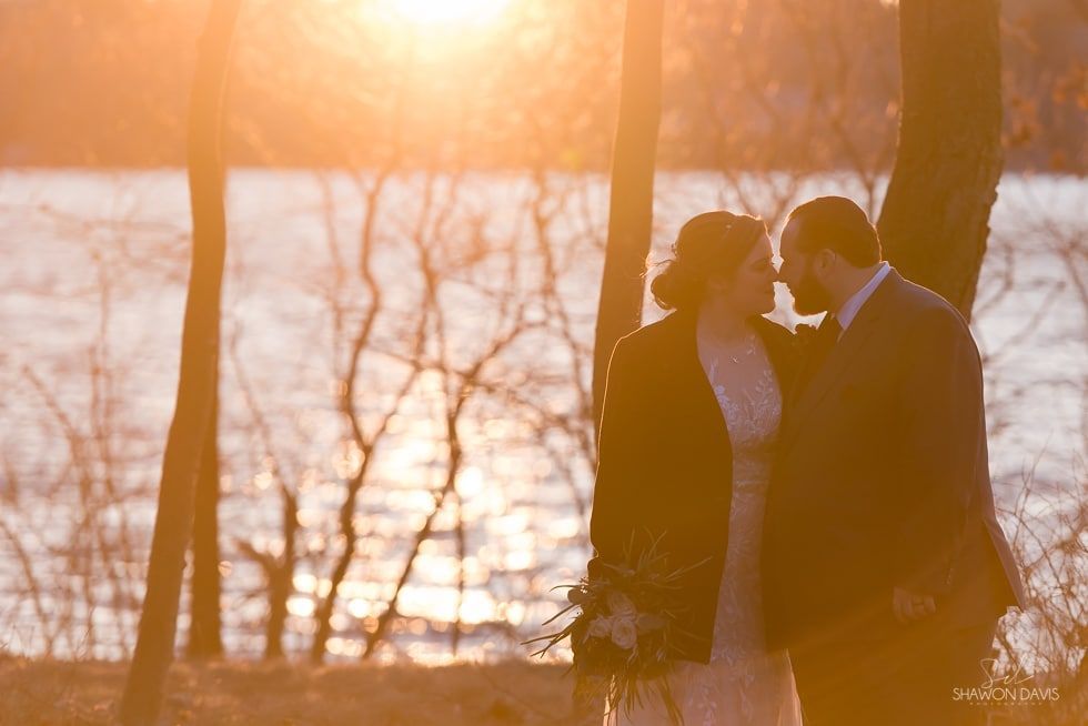 Two people in formal attire embracing by a lakeside at sunset, with warm orange light.
