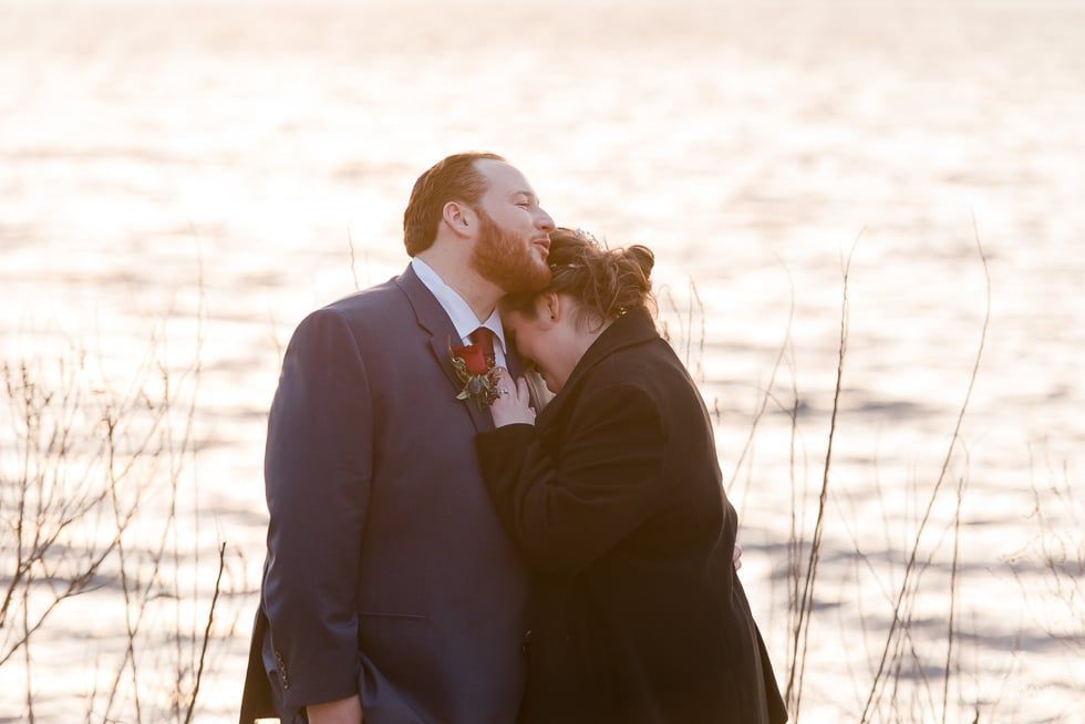 Two people embracing by a bright lakeshore at sunset, one in a dark coat and suit jacket.